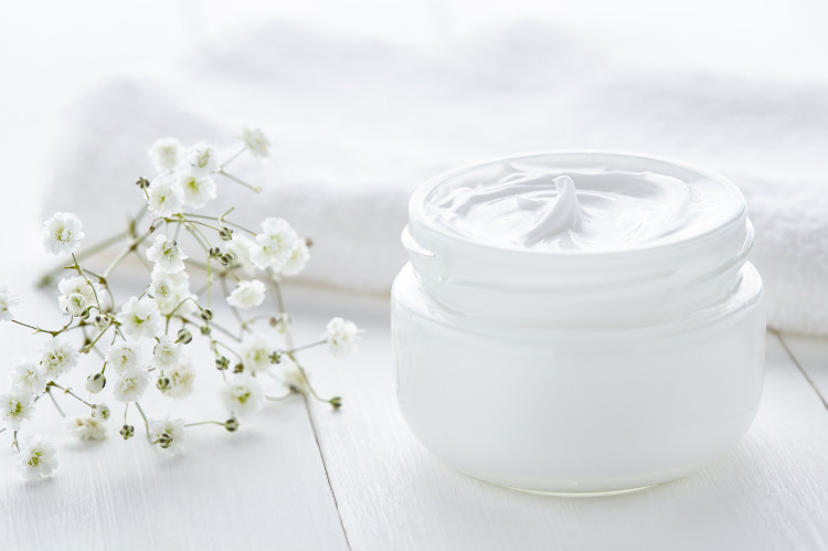 a jar of white cream on the pavement, next to a white towel and white flowers