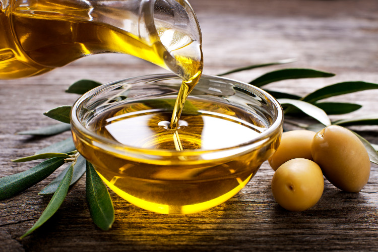 someone pours olive oil from a goblet into a bowl, next to it are olives and leaves from an olive tree