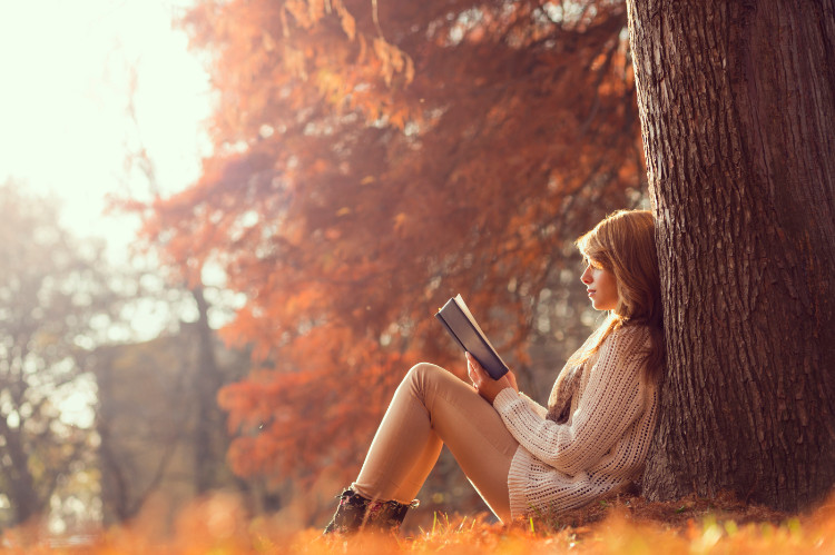woman sitting in the forest, leaning against a tree reading a book in autumn