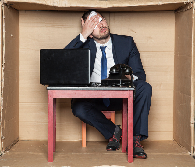 Businessman sitting at a desk in a box, cramped quarters, work stress and increased workload, computer and phone, man wiping his brow
