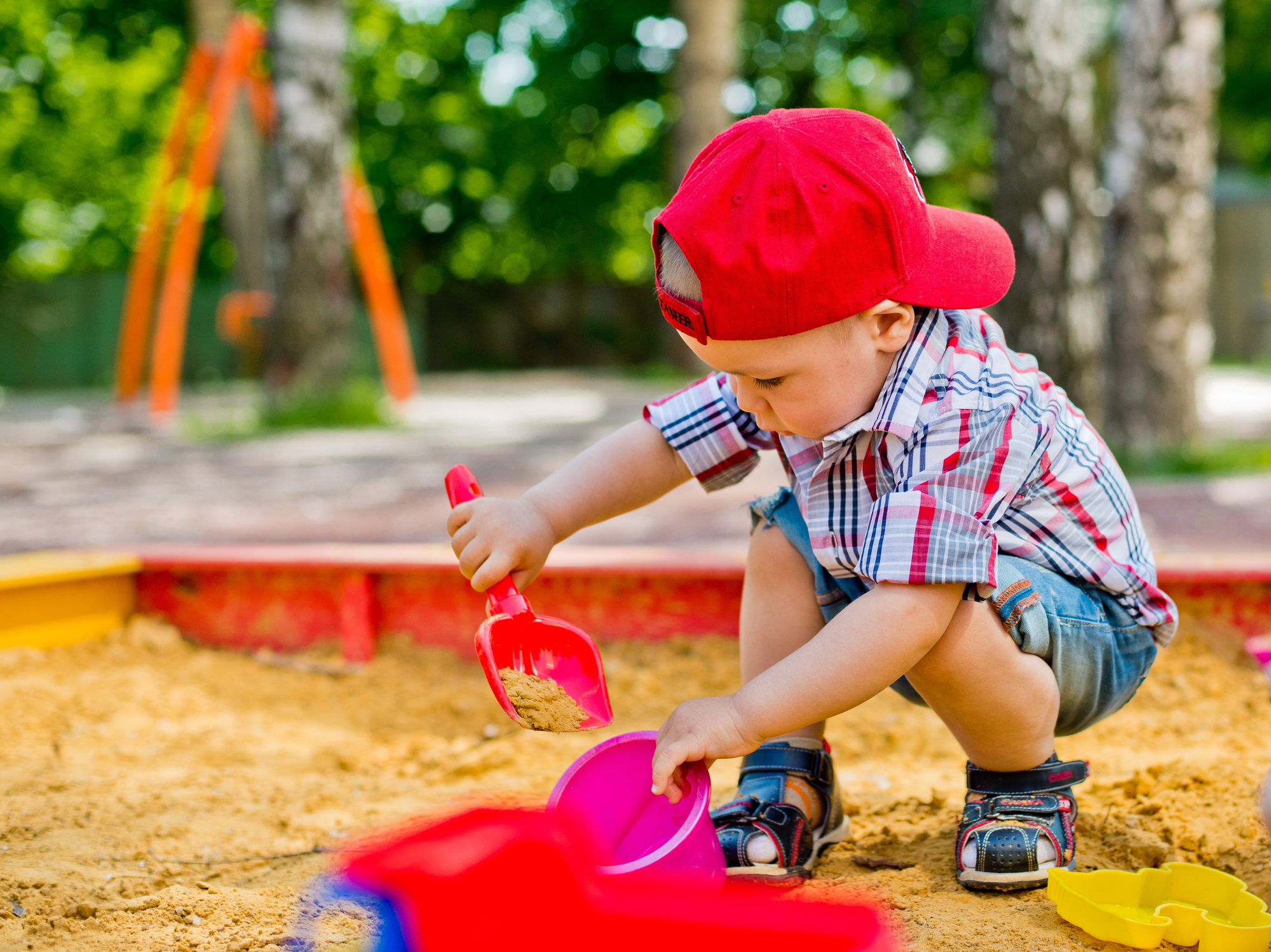 A boy scooping sand with a shovel into a bucket in a sandbox.