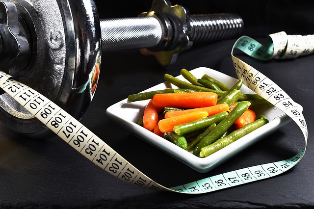 A chrome dumbbell placed next to a square plate. On the plate is a vegetable. Next to it is a tailor's tape measure.