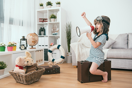 A little girl in a sheepskin coat sitting on a suitcase in the living room holding a megaphone