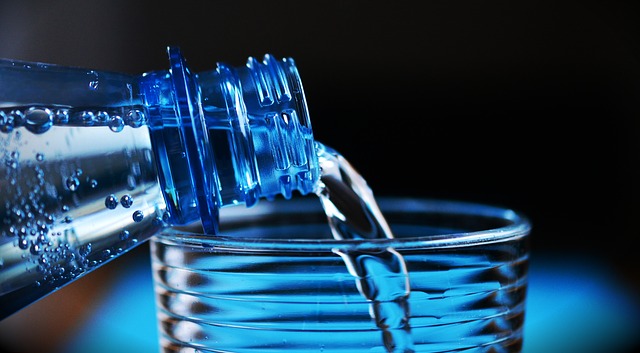 Water is poured from the bottle into the glass. Only the neck of the bottle and the top of the glass are visible. The background is dark and blue.