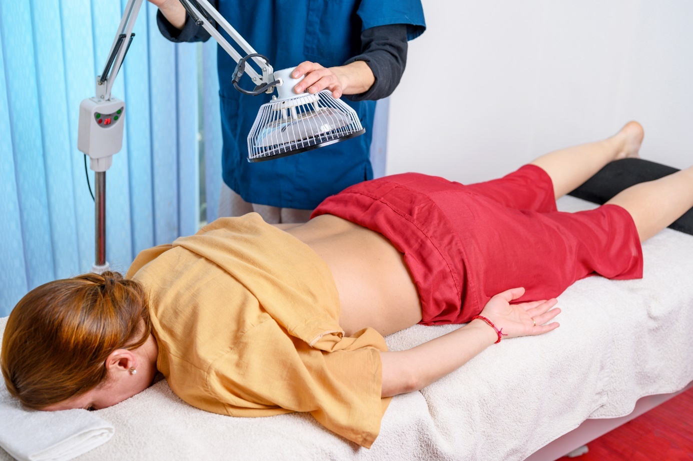 A woman is lying on a bed and a man is placing a biolamp on her sacral area.