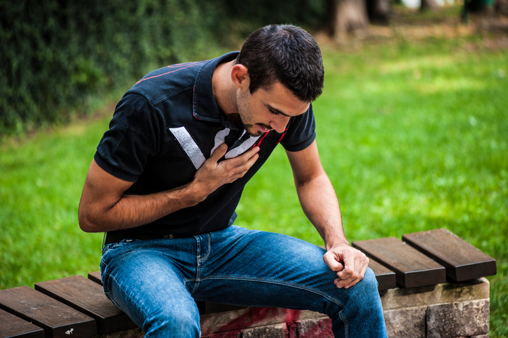 Young man sitting on park bench, holding his chest, chest pain, difficulty breathing