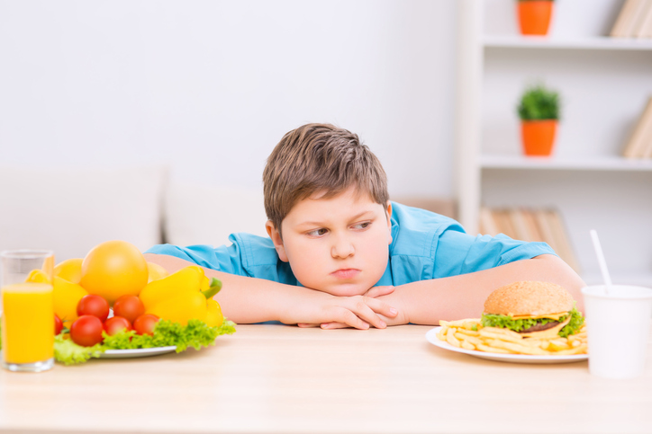 A fat kid between two plates of healthy and unhealthy food.