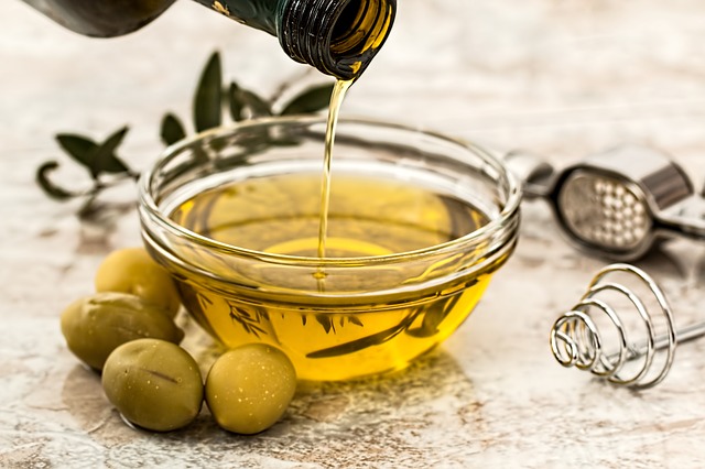 Olive oil is poured into a glass bowl from a glass bottle. Green olives are placed around the bowl.