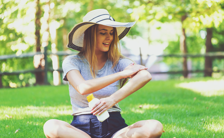 A young woman puts on sunscreen, sitting in a meadow, wearing a hat to protect her head