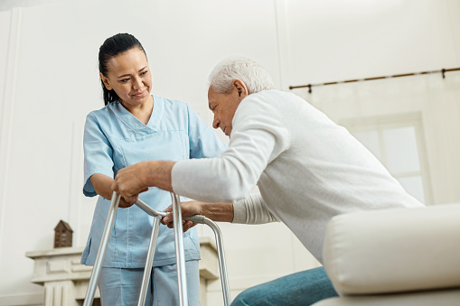 an elderly man sitting on a hospital bed trying to stand up with the help of a walker and a nurse