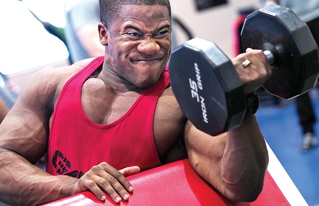 Man in red tank top. He's doing biceps with a one-arm barbell.