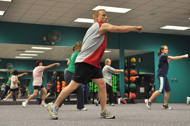 There are five people working out in the gym. One man and four women. They're dressed athletically, with their backs to a large mirror. The wall is green and the carpet on the floor is grey.