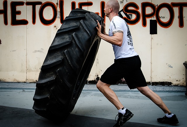 A man in a white T-shirt and black shorts is training. He's flipping a big tractor tire cover in front of him.