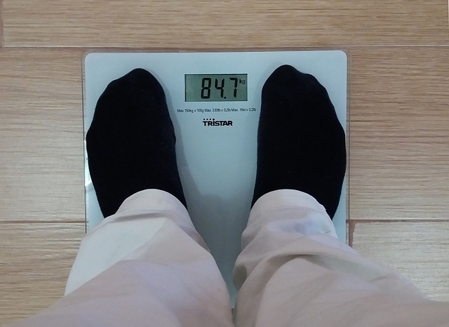 A man wearing light-coloured trousers and black socks is standing on a digital scale. He is measuring his weight. The dial reads 84.7 kilograms. The scale is white, glass, on a light-coloured floating floor.