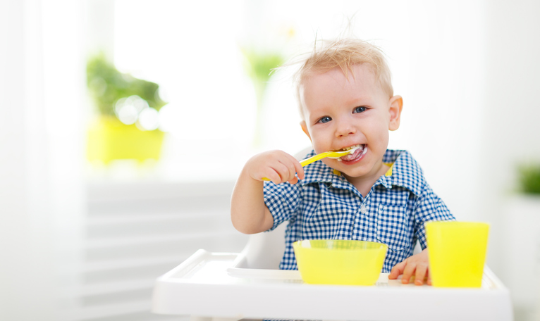 A small child sitting in a baby seat, feeding himself. Yellow spoon, cup and bowl