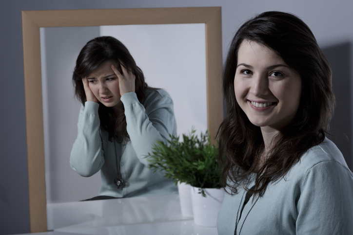 Girl has affective disorder (bipolar affective disorder) or cyclothymia, standing in front of mirror laughing, manic episode and in mirror sad, bad mood, depressive episode