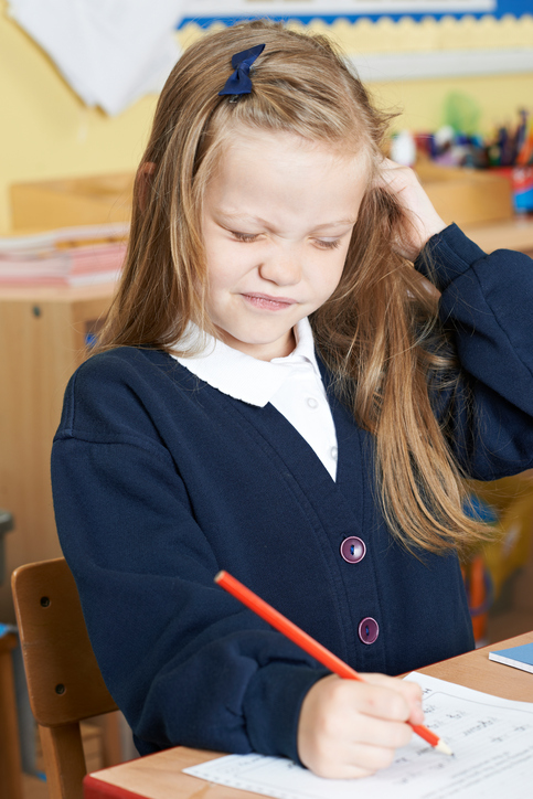 A little girl sitting at school with an itchy head, probably lice in her hair, blonde hair, sitting and writing