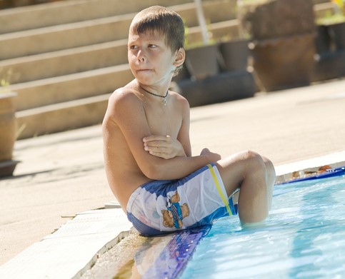 The boy is sitting by the pool, his feet in the water. He's cold.